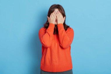Güzel, koyu renk saçlı bir kadın yüzünü avuç içiyle kapatıyor, gözlerini kapatıyor, esmer kız turuncu kazak giyiyor, mavi arka planda yalnız poz veriyor, birinden saklanıyor..