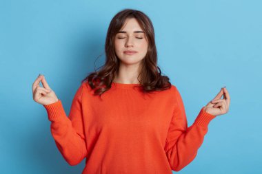 Genç, güzel bir kadın meditasyon yapıyor, iki elini de iyi bir jestte tutuyor, ve gözleri kapalı duruyor, rahatlamak için yoga yapıyor, turuncu kazak giyiyor, mavi arka planda izole edilmiş..
