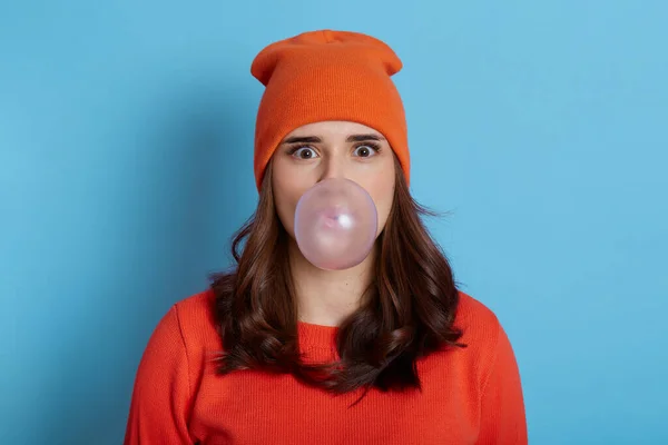 Young pretty lady with big eyes holding big chewing gum inside mouth, blowing large bubble, wearing cap and sweater, posing isolated over blue background.