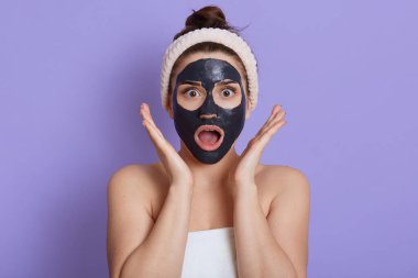 Nutku tutulmuş, ağzı açık, çamur maskesi takan, güzellik prosedürleri uygulayan, şok olmuş yüzü olan, vücuduna havlu sarılmış, leylak duvarda izole edilmiş, avuç içlerini yüzüne yakın tutan bir kadının fotoğrafı..