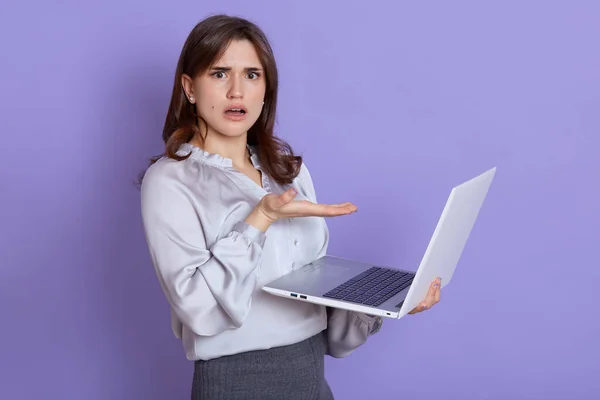 Young European woman frustrated by computer operational problems, looking at camera with puzzled and astonished expression, has unexpected troubles, pointing at laptop screen with palm.