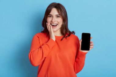 Koyu renk saçlı kadın boş akıllı telefon görüntüsü veriyor, komik bir mesaj aldığı için mutlu, internette sörf yapıyor, turuncu kazak giyiyor, ağzını açık tutuyor ve yanağına dokunuyor, bağırıyor, mutlu bir şekilde..