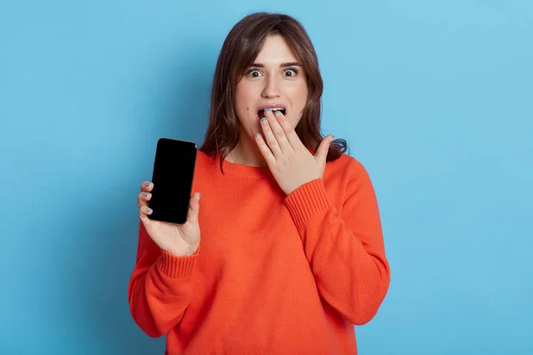 Wanita Berambut Gelap Yang Terkejut Memakai Sweater Oranye Memegang Ponsel — Stok Foto