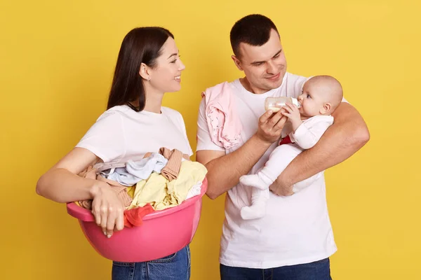 洗濯や哺乳瓶からの新生児の女の子の給餌を行う幸せな両親 母は洗濯のためのリネンをベースに保持しています 白いTシャツを着てお母さんとお父さん 黄色の背景に隔離された立って — ストック写真