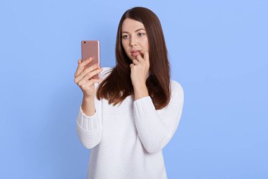 Düşünceli siyah saçlı kadın düşünceli akıllı bir telefon ekranı gibi görünüyor, cep telefonu tutuyor, beyaz kazak giyiyor, işaret parmağını çenesinde tutuyor, mavi duvara karşı içeride poz veriyor..