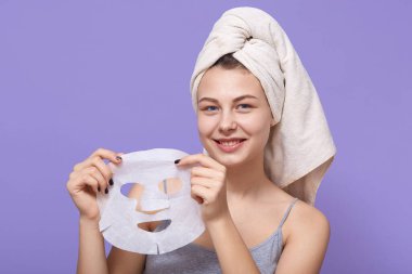 Hoş görünümlü, havluya sarılı, gri kolsuz tişörtlü, elinde doku kozmetik maskesi olan, kameraya güzellik ürünlerini gösteren ve leylak duvara gülümseyen çekici bir kadın..