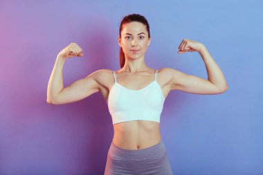 Genç ve güçlü bir kadın doğrudan kameraya bakıyor, iki elini kaldırıyor ve pazılarını gösteriyor, üst ve tozluklarını giyiyor, renkli arka planda soyutlanmış, mükemmel vücutlu ve at kuyruklu bir kadın..