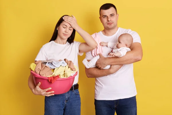 Tired parents doing household chores and taking care of newborn baby, mother holds basin with linen, keeps eyes closed and touch forehead with palm, father carries daughter with exhausted expression.