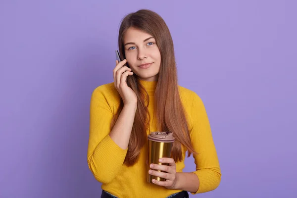 Positive female talks with interest and joy via cellular, wears casual attire, enjoys drinking aromatic coffee fro thermo mug, isolated over lila background, lady looks at camera.
