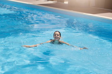 Havuzda rahatlayan, yüzme havuzunda gülümseyen, neşeli bir yüz ifadesiyle kameraya bakan, lüks bir otelde dinlenirken mutluluğu hisseden bir kadın..