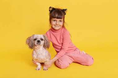 Koyu renk saçlı, köpek yavrusu gibi poz veren küçük bir kız, kameraya bakan ve gülümseyen, hayvanına sarılan, sarı arka planda izole edilmiş kedinin kulaklığı ve günlük kıyafet giyen bir çocuk..