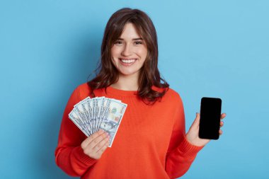 Portakal rengi günlük giysiler içinde mutlu bir genç kadın, elinde cep telefonu, elinde nakit para vantilatörü, mavi duvarda izole edilmiş, kameraya bakıyor, neşe ifade ediyor..