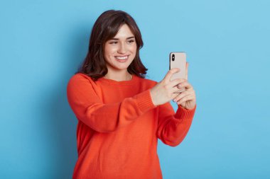 Genç ve çekici bir kadının portresi. Mavi arka planda izole edilmiş akıllı bir telefonla selfie çekiyor. Kadın mutlu bir gülümsemeyle cihaza bakıyor..