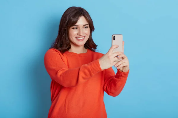 Portrait of young attractive woman making selfie photo with smart phone isolated over blue background, lady looks at device with happy smile, dark haired female has video call.