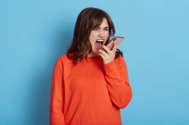 Kızgın kadın telefonda konuşurken bağırıyor, çok kızgın, turuncu günlük kıyafetler giyiyor, mavi arka planda tek başına duruyor, biriyle tartışıyor, hoş olmayan bilgiler veriyor, kızıyor..