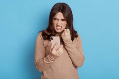 Öfkeli sıradan bir kadın mavi arka planda izole edilmiş akıllı bir telefon kullanıyor, suratını asıyor ve yumruğunu öfkeyle sıkıyor, vitrine bakıyor, dişlerini gösteriyor, yatak haberlerini görüyor, günlük giysiler giyiyor..