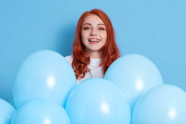 Neşeli kızıl saçlı kadın çok mutlu ve heyecanlı, boş zamanlarını partiye harcıyor, beyaz gömlek giyiyor, mavi duvarda izole edilmiş balonların yanında poz veriyor, heyecanlı kızıl kız kameraya bakıyor..