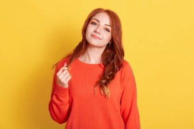 kızıl saçlı Avrupalı model, hoş görünüşlü, neşeli gülümseme, sakin yüz ifadesi, uzun kollu turuncu kazak giyiyor, sarı arka planda duruyor, saçına dokunuyor ve doğrudan kameraya bakıyor..