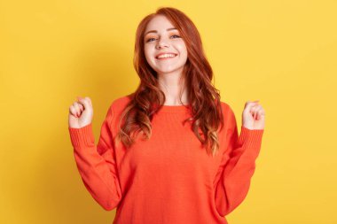 Aşırı neşeli kızıl saçlı, büyüleyici gülüşlü, ellerini yumruk yapıp kaldıran, uzun zamandır beklenen tatillerden hoşlanan, günlük turuncu kazak giyen, güzel duygular ifade eden, sarıda izole edilmiş bir kadın.