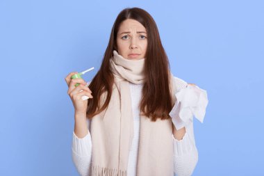 Çekici, hasta, koyu renk saçlı kadın boğaz ağrısını gidermek için sprey kullanmak istemiyor. Yorgun görünüyor ve kötü hissediyor. Mavi arka planda yalnız poz veriyor. Corona virüsü, sağlık sorunları.