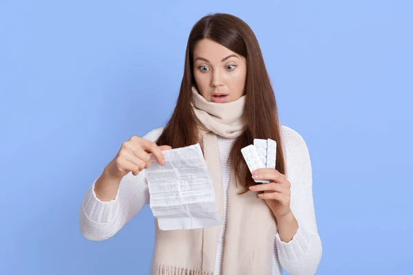 Close up portrait of disappointed sad lady holding pills and its instruction in hands, thinking about taking drugs, looking at paper with widely opened mouth and shocked facial expression.