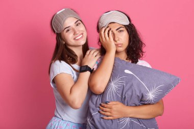 Üzgün kıvırcık kadın üzgün bir şekilde kameraya bakar ve yüzünün yarısını avuç içi ile kaplar, arkadaşı yanında mutlu bir şekilde durur ve gülümser, pijama giyer ve gözbağı takar, pembe arka planda durur..