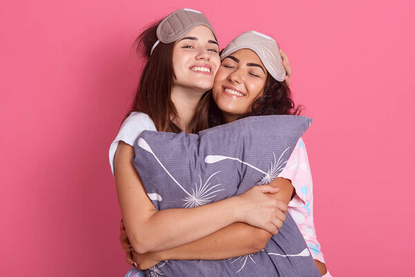 Две счастливые темноволосые женщины в пижамах и повязках на глаза, обнимая и обнимая подушку, закрывая глаза, выражая счастье, наслаждаясь проведением времени вместе.