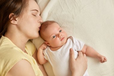 Uykulu, uykulu, gözleri kapalı bir anne yeni doğmuş bebeğiyle yatağa uzanıyor ve bebeği öpüyor, sevimli bir çocuk, dış görünüşte çalışıyor, beyaz bir beden giyiyor..