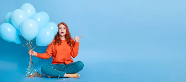 Young ginger astonished female sitting on floor and pointing aside with thumb, holds balloons, keeps mouth opened, sees something shocking, woman posing against blue wall, copy space for advertisement