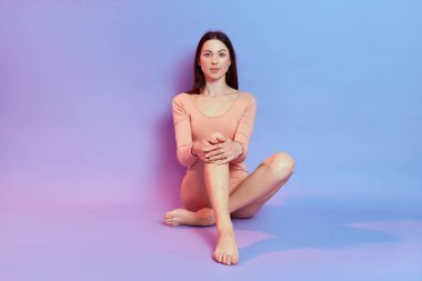 Koyu renk saçlı, mavi arka planda bej bir elbise giyen, pembe ışıklı, doğrudan kameraya bakan, kendine güvenen, elini dizinde tutan, yalın ayak bir kadın..