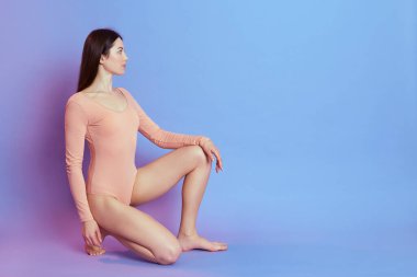 Çekici, tapılası, bej renkli bir elbise giyen, dizlerinin üstüne çöküp başka tarafa bakan, kendine güvenen ve mükemmel bir vücudu olan, mavi arka planda izole edilmiş bir kadın. Promosyon metni için boşluk kopyala.