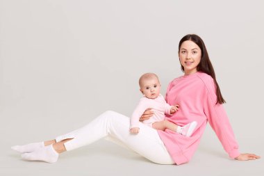 Genç Avrupalı anne kucağında küçük kızıyla yerde oturuyor, annesinin bacaklarında bebek tulumu, beyaz pantolon ve beyaz duvarda bebekle poz veren pembe kazak giyen bir kadın..