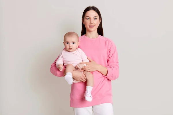 Closeup portrait of beautiful dark haired woman wearing pink sweatshirt and pants holding on hands her little daughter on white background. Family, love, lifestyle, motherhood and tender moments.
