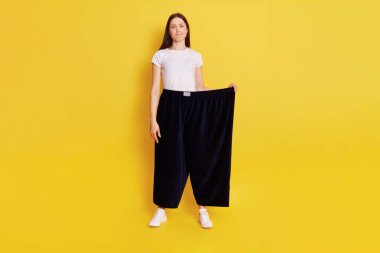 Full length photo of Young adult Caucasian girl wearing casual t shirt and too big size pants, looks at camera with puzzled expression and bewilderment, purses lips, isolated over yellow background. clipart