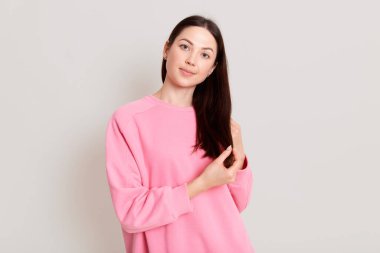 Pembe kazak giyen çekici, koyu renk saçlı bir kadının portresi. Sakin bir ifadeyle kameraya bakıyor ve gülümsüyor. Elleriyle saçlarına dokunuyor..