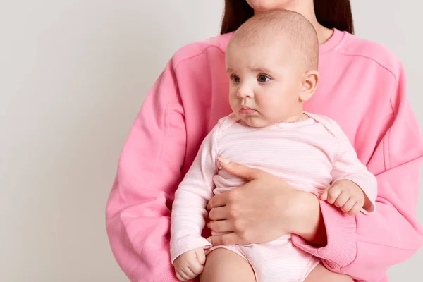 Profile of dark haired female wearing casual pink sweatshirt and pants sitting on floor with cross legs and holds hands of her little daughter, infant dresses bodysuit learns to stand with mother help