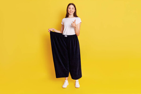 Attractive dark haired female wearing old big trousers after weight lost, has happy, proud expression, pointing index finger to camera, motivates to grow thin, isolated over yellow background.