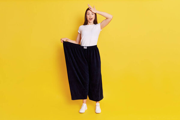Full length photo of surprised female dresses huge size trousers, looking at camera with shocking expression and touching her head with astonishment, isolated over yellow background.