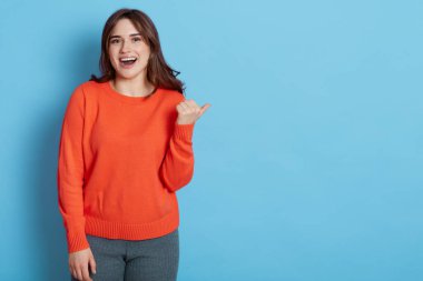 Turuncu süveter giyen heyecanlı kadın mutlu bir yüz ifadesini işaret ediyor, hoş koyu saçlı ve dişlek gülüşlü, mavi arka planda soyutlanmış bir kadın..