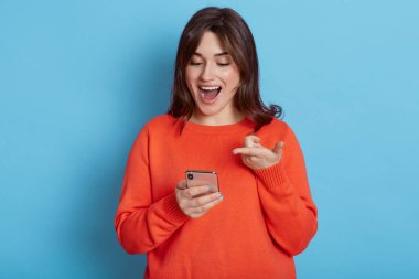 Mutlu, koyu renk saçlı, günlük kıyafetler giyen, elinde akıllı bir telefonla duran, ağzı açık ve heyecanlı bir ifadeye sahip, mavi arka planda izole edilmiş bir cihaz ekranına bakan Avrupalı kadın..