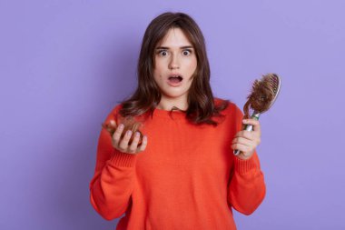 Şok olmuş yüz ifadesi olan güzel bir kadın, saç dökülmesinden dolayı üzgün, tedaviye ihtiyacı var, üzerinde bir sürü saç olan tarak tutuyor, kazak giyiyor, açık ağzıyla leylak arkaplan üzerinde duruyor..