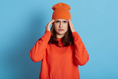 Baş ağrısı çeken, şakaklara iki eliyle dokunan, migreni olan, günlük turuncu kazak ve şapka giyen, tedaviye, sağlık hizmetine ihtiyacı olan, mavi arka planda yalnız poz veren bir kadın..