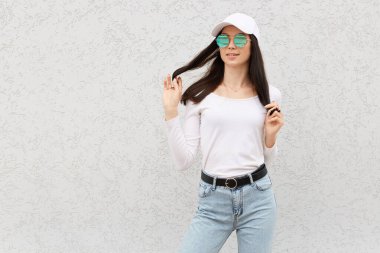 Kendine güvenen güzel bir kadın ayakta duruyor ve başka tarafa bakıyor, pozitif duygularını ifade ediyor, mutlu bir şekilde gülüyor, kot pantolon giyiyor, rahat bir gömlek ve beyzbol şapkası giyiyor, aydınlık duvarın üzerinde izole edilmiş..