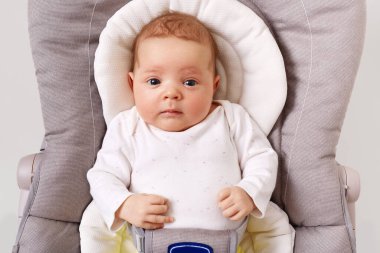Front view of curious new born baby wearing white podysuit lying in child bouncer chair, looking at camera, exploring world, little infant, happy childhood.