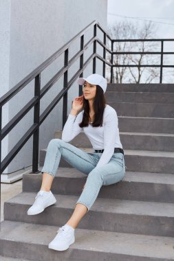 Attractive female wearing stylish jeans, white shirt and baseball cap, sitting on stairs and looking away with thoughtful expression, posing outdoor near building.