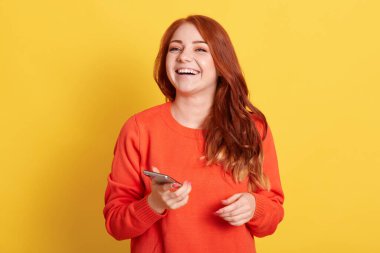 Happy ginger female wearing casual orange sweater, holding smart phone in hands and laughing happily, looking at camera with positive emotion, isolated over yellow background.