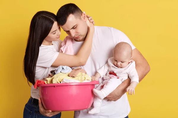 快乐疲惫的家庭 深色头发的女人拥抱着他的丈夫 男人摆姿势抱着新生的孩子 满满的衣服 疲惫的父亲做完家务活 照顾好孩子 — 图库照片