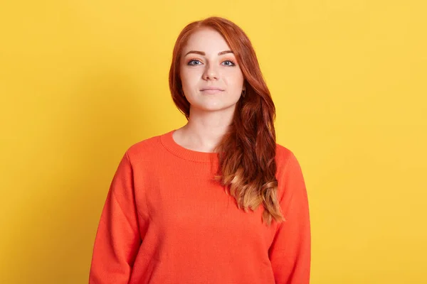 stock image Front view of serious young attractive woman with red hair wearing casual orange sweater, looking directly at camera with confident expression, isolated over yellow background.