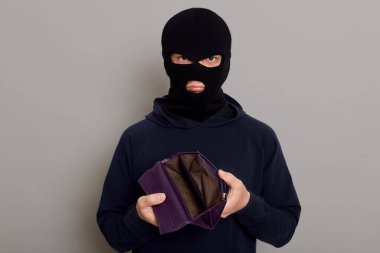 Upset burglar dressed in black turtleneck and balaclava holding empty wallet, feeling frustrated, looking at camera with pouting lips, being unhappy with theft, isolated over gray background.