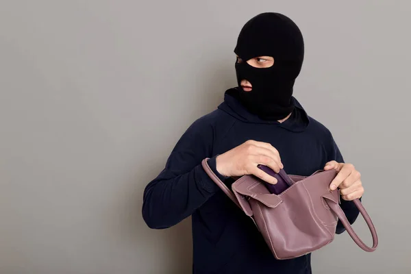 Insidious male villain in balaclava stands with stolen women's bag and wallet, afraid that he will be caught, turns around and looks carefully back, isolated over gray background.